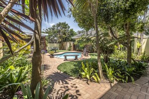 Un jardín con piscina, un banco de piedra y diversas plantas.