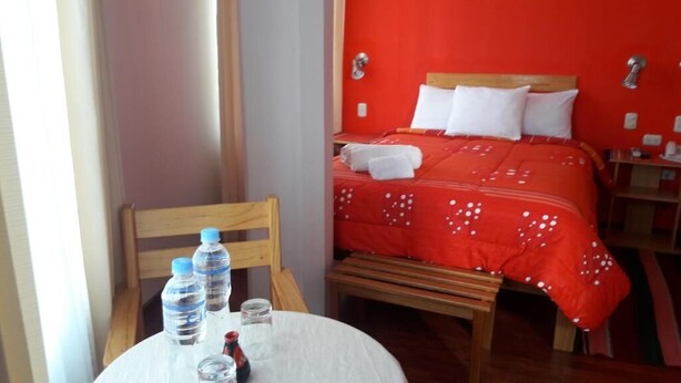 A hotel room with a bed, a wooden chair, a table with water bottles, and a red wall. - Kuntur Inn (Puno)