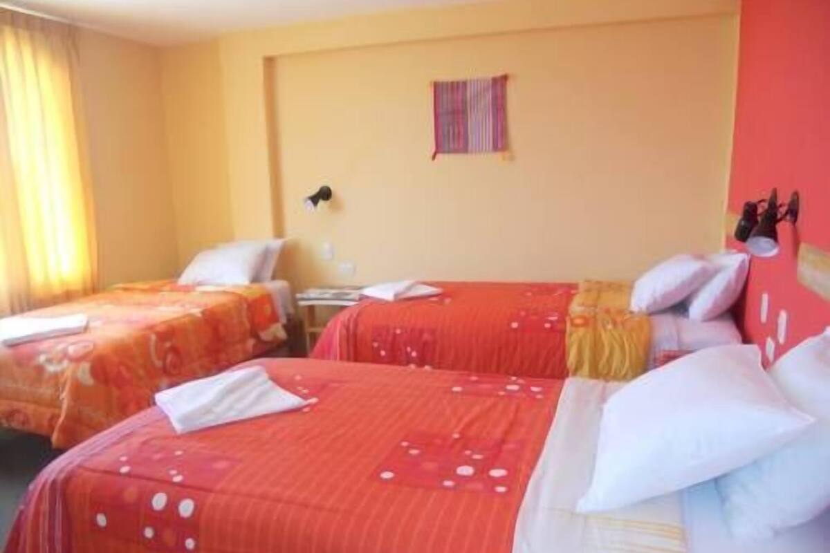 A hotel room with two beds, each with red bedding and white pillows. There is a small table and a wall-mounted lamp.