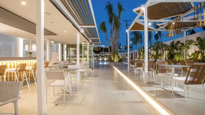 An outdoor cafe area with tables and chairs, palm trees, and a modern architectural design. - Casa Stavris (Rhodes)