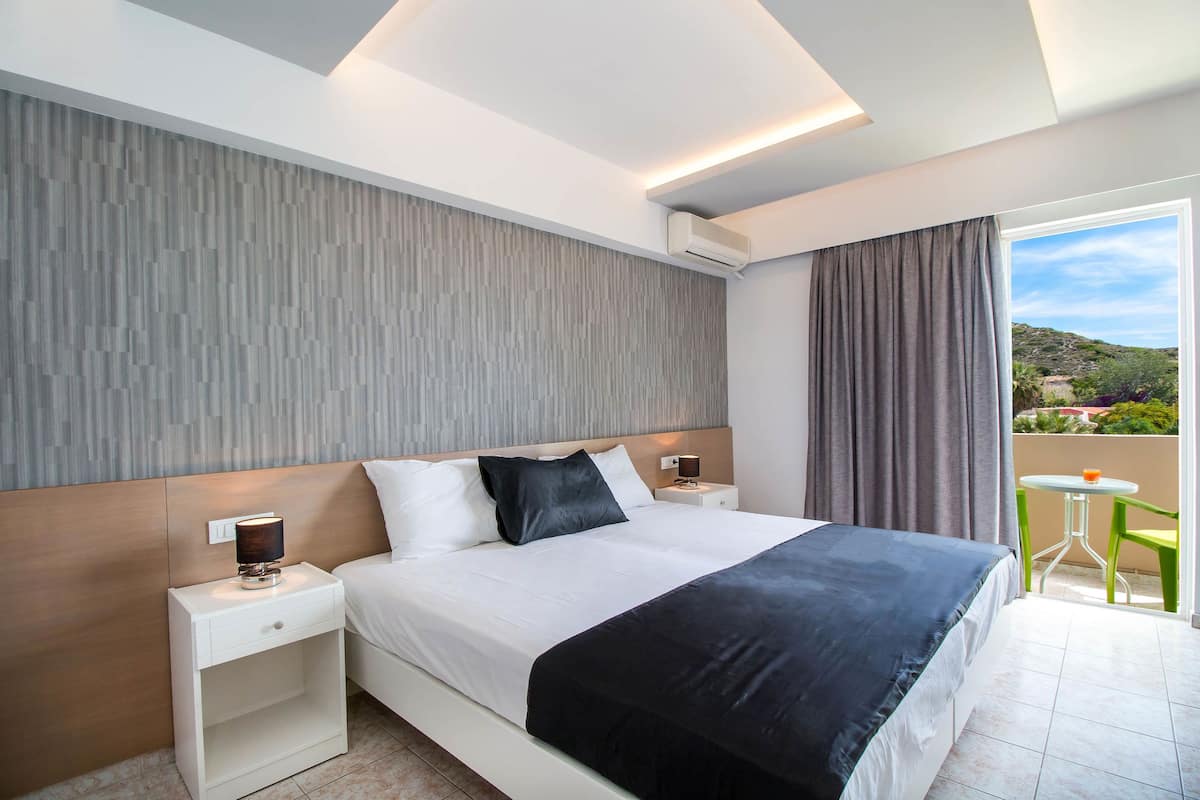 Una habitación de hotel moderna con una cama grande, una mesita de noche con lámpara, una pequeña mesa con silla y vistas al exterior.