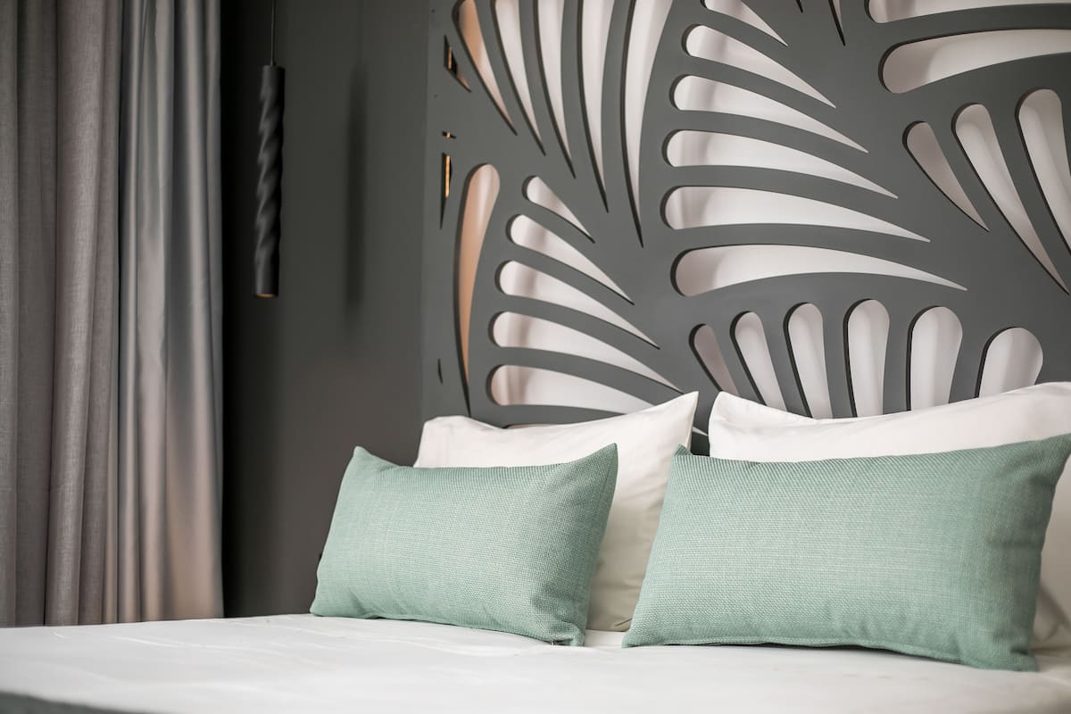 Una cama con almohadas blancas y verdes, una cortina gris y una pared con un diseño de hojas.