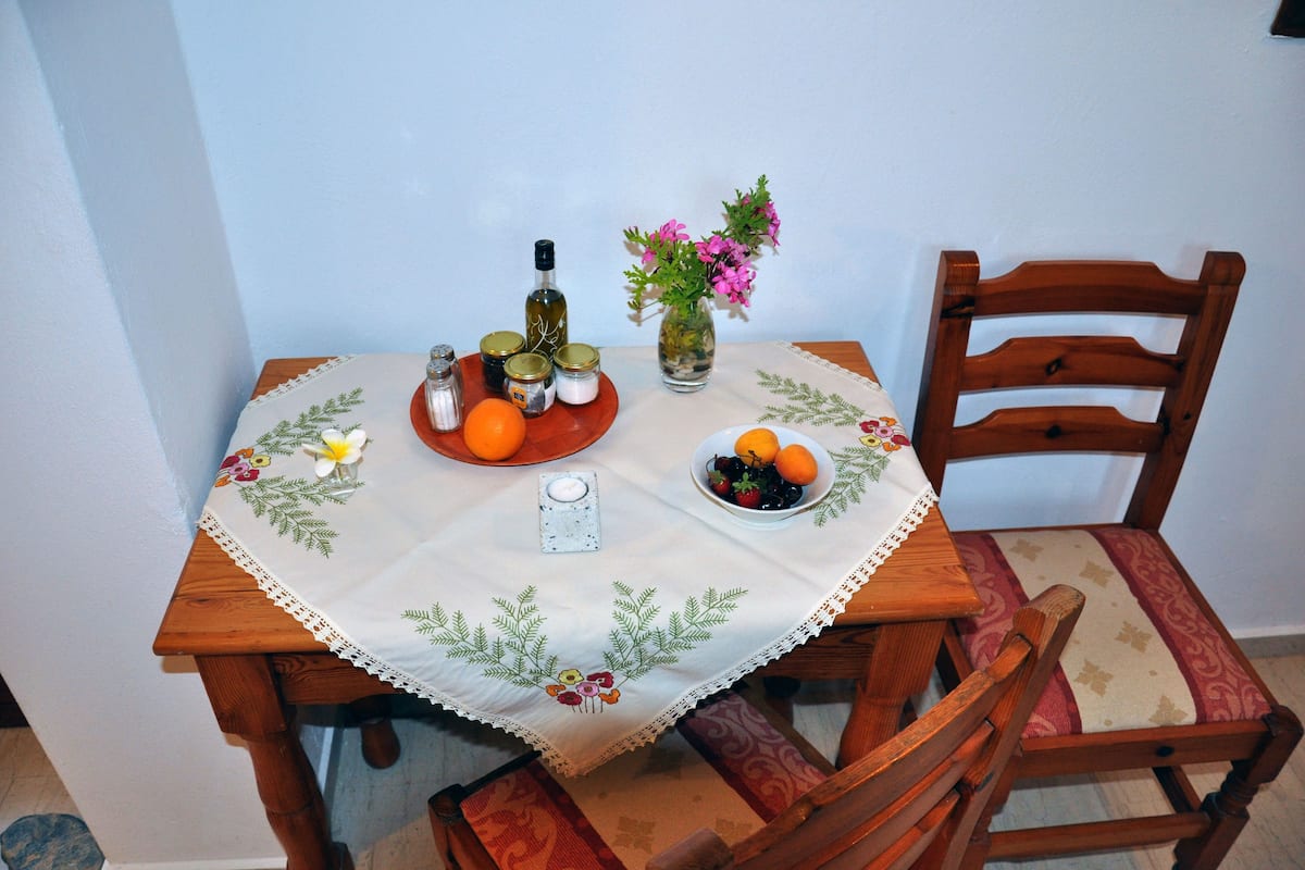 A wooden dining table with a white tablecloth, a vase with flowers, a bowl of fruit, and a bottle of olive oil.