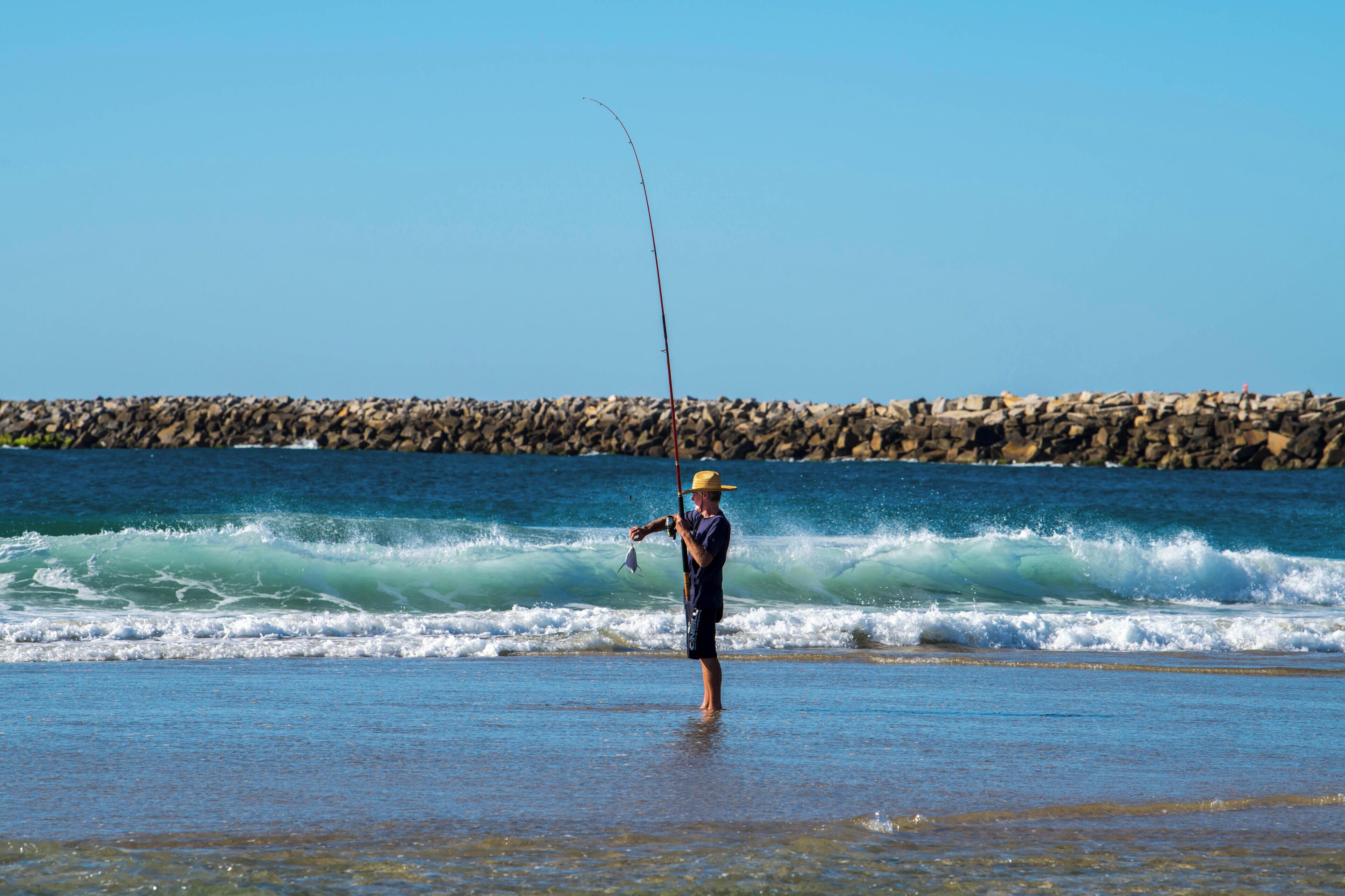 A person standing in shallow water near a rocky shoreline, holding a fishing rod.