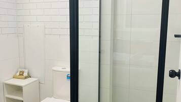 A bathroom with a white toilet, a small shelf with toiletries, and a glass shower enclosure.