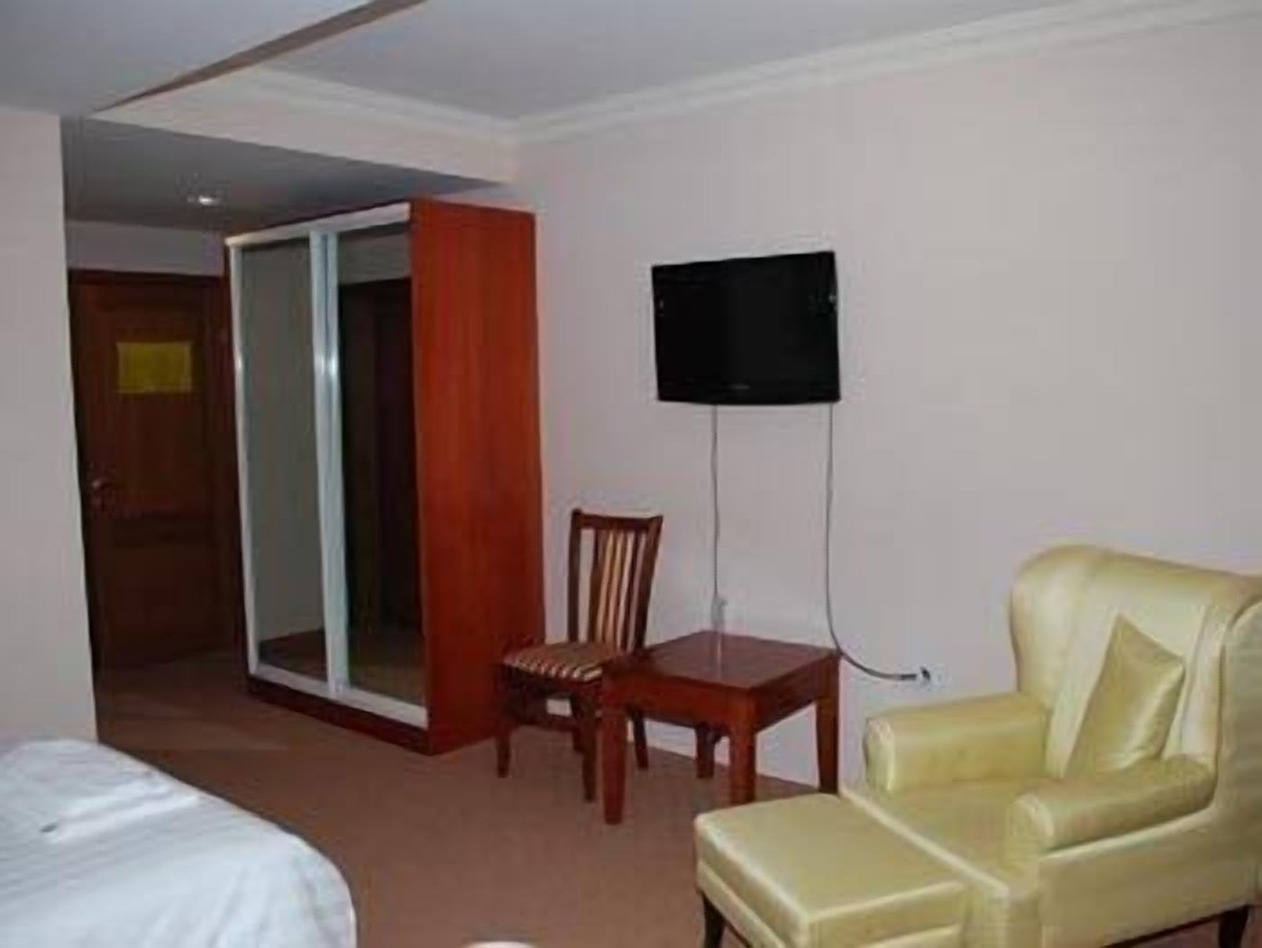A hotel room with a bed, a wooden chair, a yellow armchair, a television mounted on the wall, and a sliding door.