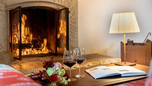 A fireplace with a fire, a table with a book and wine glasses, and a lamp. - Castello di Petrata (Assisi)