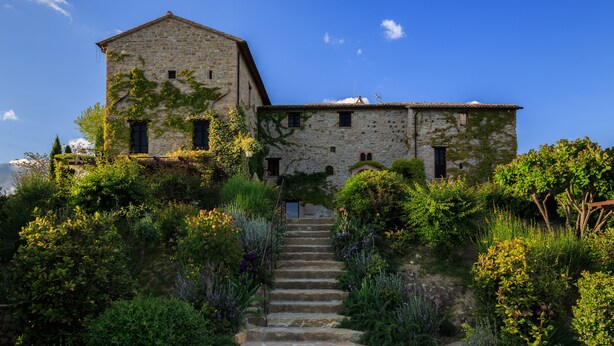 A stone building with a staircase leading to a door, surrounded by lush greenery. - Castello di Petrata (Assisi)