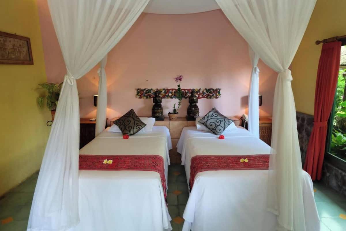 A room with two beds, each with white linens and red decorative borders. There is a canopy over the beds, and a wall-mounted shelf with decorative items.