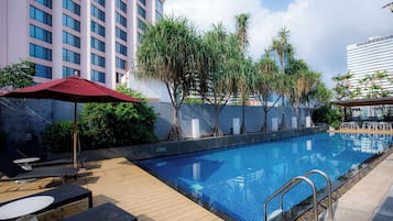 Piscina com espreguiçadeiras e guarda-sóis, cercada por árvores e edifícios modernos.