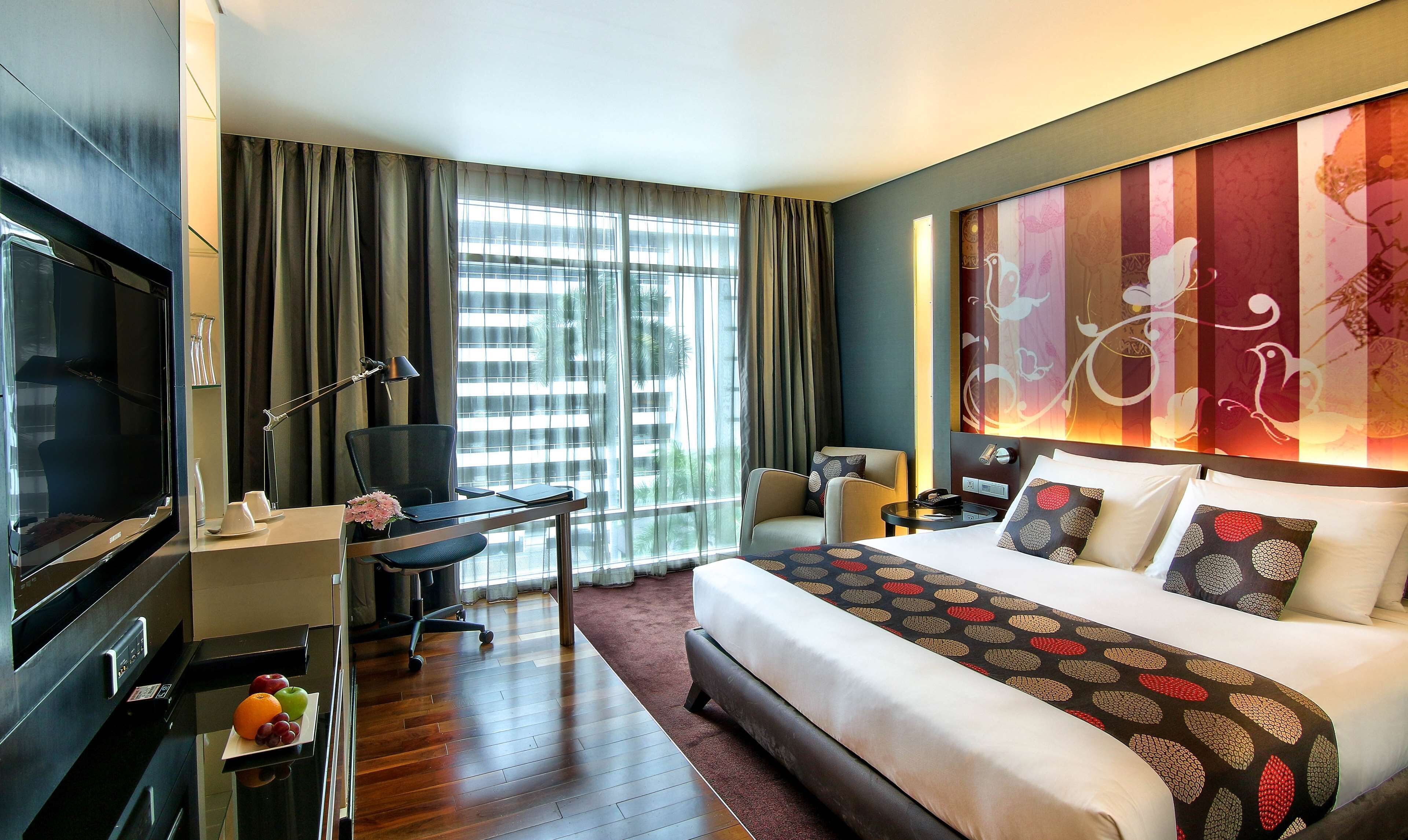 A hotel room with a large bed, a desk with a chair, a television, and a view of the city through large windows.