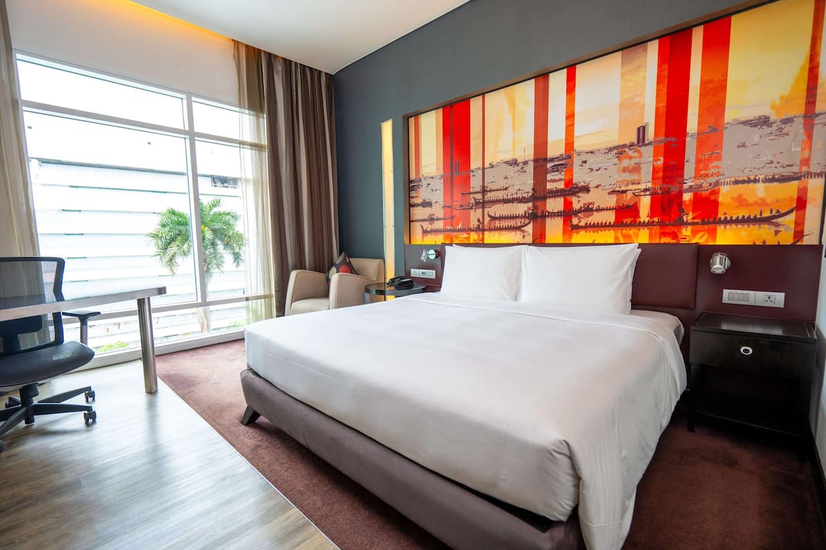 A hotel room with a large bed, a desk, and a chair. There is a window with curtains, a painting on the wall, and a plant outside.