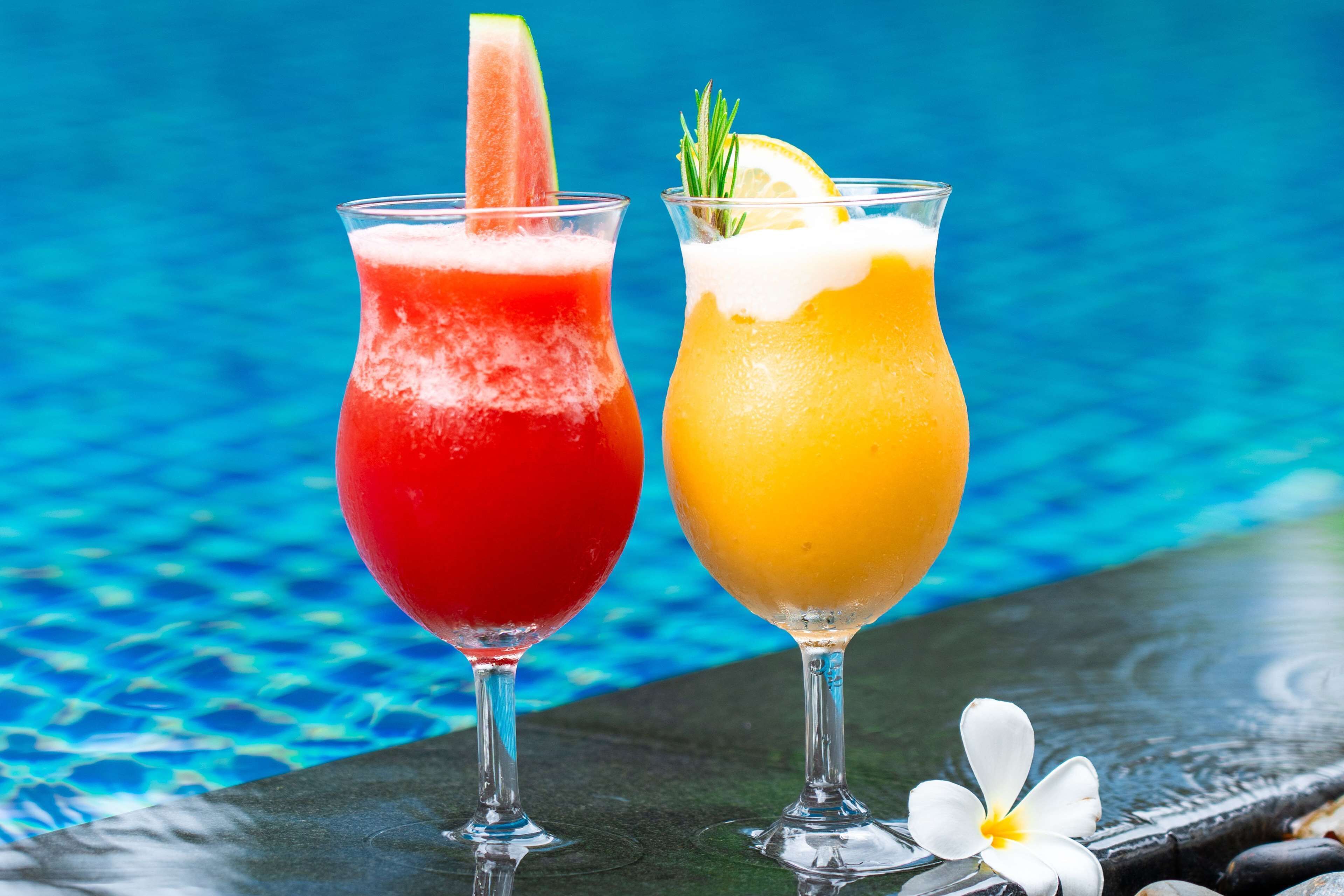 Two cocktails with garnishes, placed on a table by a pool.