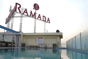 Un espace autour d’une piscine avec une grande enseigne indiquant « RAMADA » et un panneau d’affichage contenant des informations.