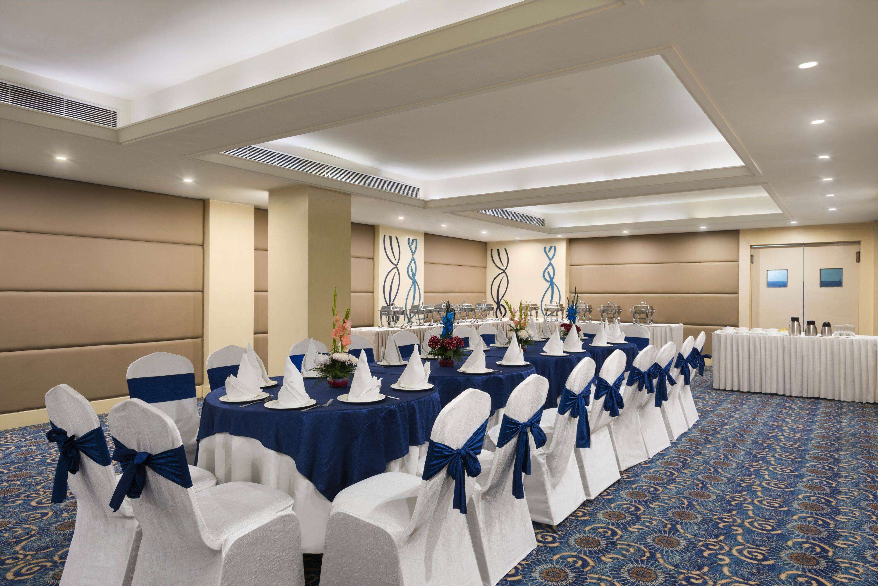 Une salle de banquet avec des rangées de tables dressées pour un événement formel, avec des chaises blanches ornées de rubans bleus et des nappes bleues.