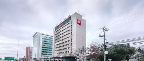 Un hotel alto con un logo rosso ben visibile, circondato da altri edifici e da una strada con auto.