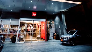 Entrada de un hotel con un llamativo letrero de 'ibis', una zona de acceso cubierta y un coche estacionado.