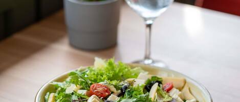 Un saladier rempli de salade, avec différentes sortes de légumes verts et diverses garnitures, un verre d’eau et une plante en pot en arrière-plan.
