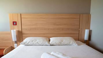 Una cama bien hecha con dos almohadas y dos toallas enrolladas, cabecera de madera y lámparas de noche.