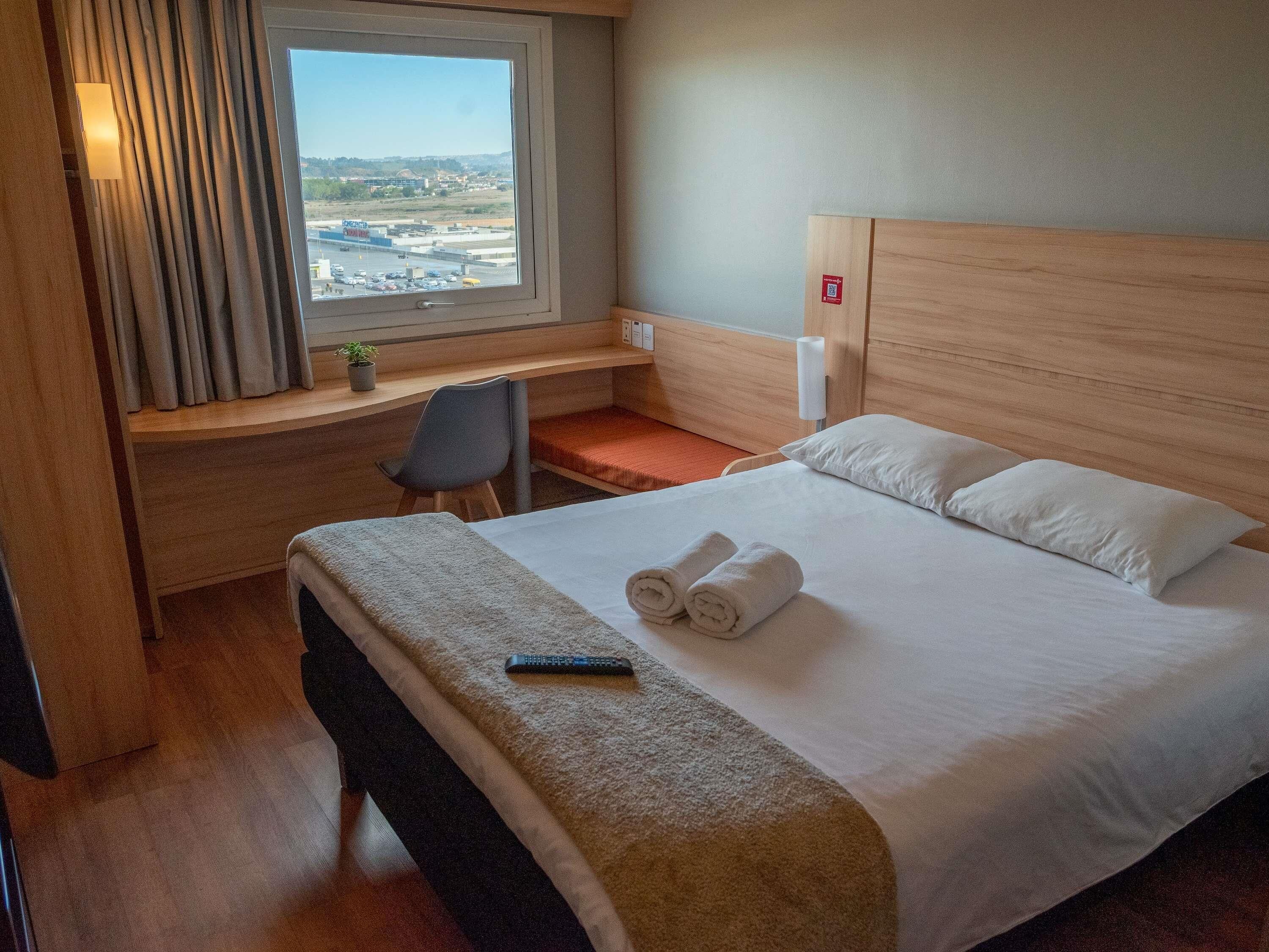 A hotel room with a bed, a desk, a chair, a window with curtains, and a view of a cityscape.