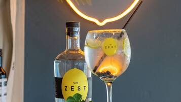 A bottle of gin and a glass with a straw, placed on a wooden surface with greenery around them, and a neon sign in the background.