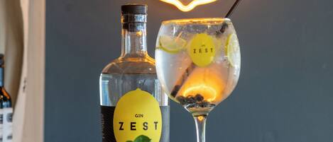 A bottle of gin and a glass with a straw, placed on a wooden surface with greenery around them, and a neon sign in the background.