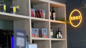 Zona de bar con un letrero de neón, estantes con botellas y aperitivos, y una máquina de café.