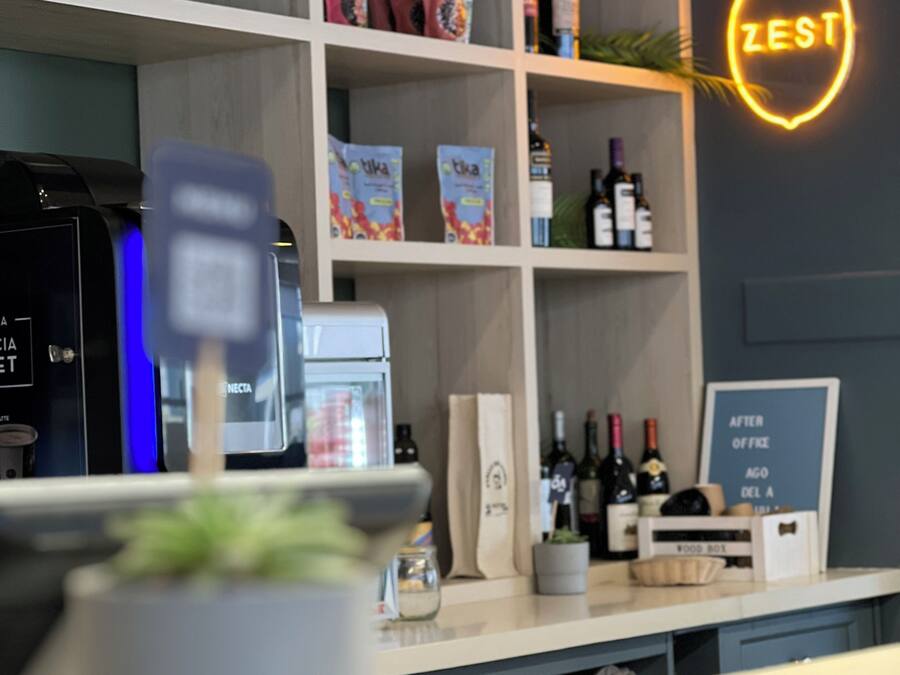 Angolo bar con insegna al neon, scaffali pieni di bottiglie e snack, e una macchina da caffè.