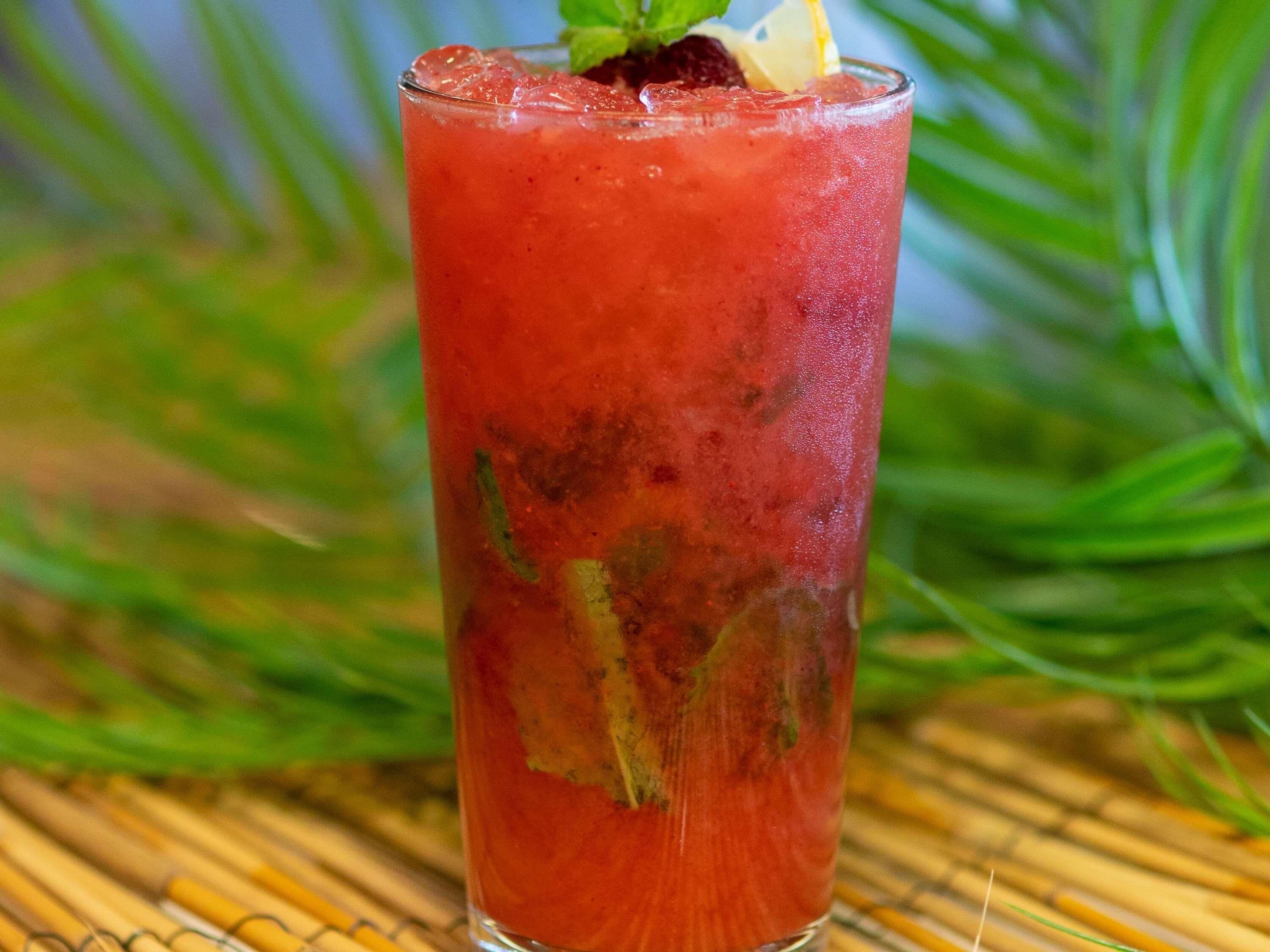 A tall glass of red beverage with a mint garnish, ice cubes, and a slice of lemon on a bamboo mat.