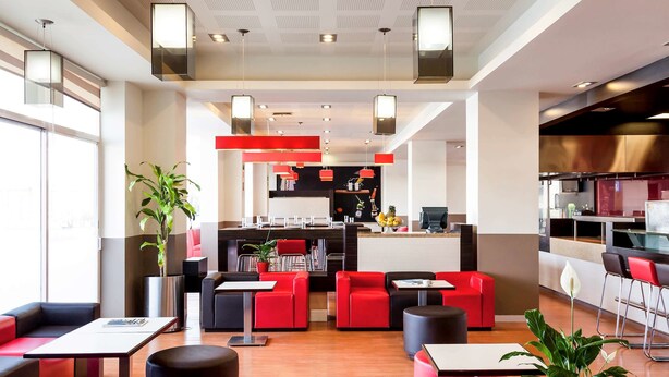 Un moderno vestíbulo de hotel con asientos rojos y negros, una isla central con zona de cocina y una gran planta en maceta.