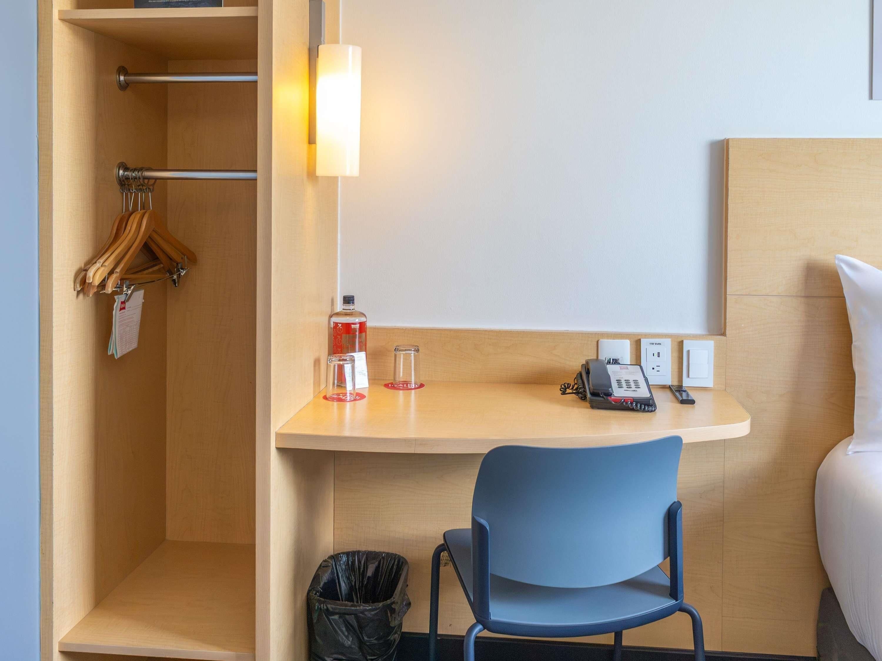 A hotel room with a wooden desk, a blue chair, a telephone, a bottle of hand sanitizer, and a trash bin.