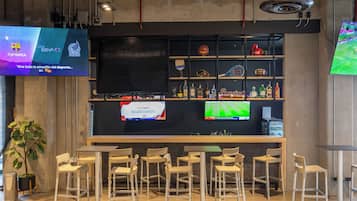 Eine Bar mit verschiedenen Sitzgelegenheiten, einem Fernseher, der Sport zeigt, und Regalen mit unterschiedlichen Artikeln.