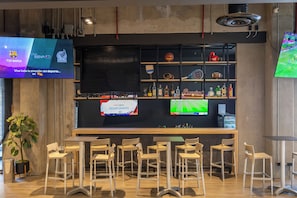 A bar area with multiple seating options, a television displaying sports, and shelves with various items.