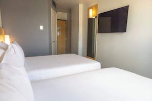 A hotel room with two single beds, a door leading to a hallway, and a wall-mounted TV.