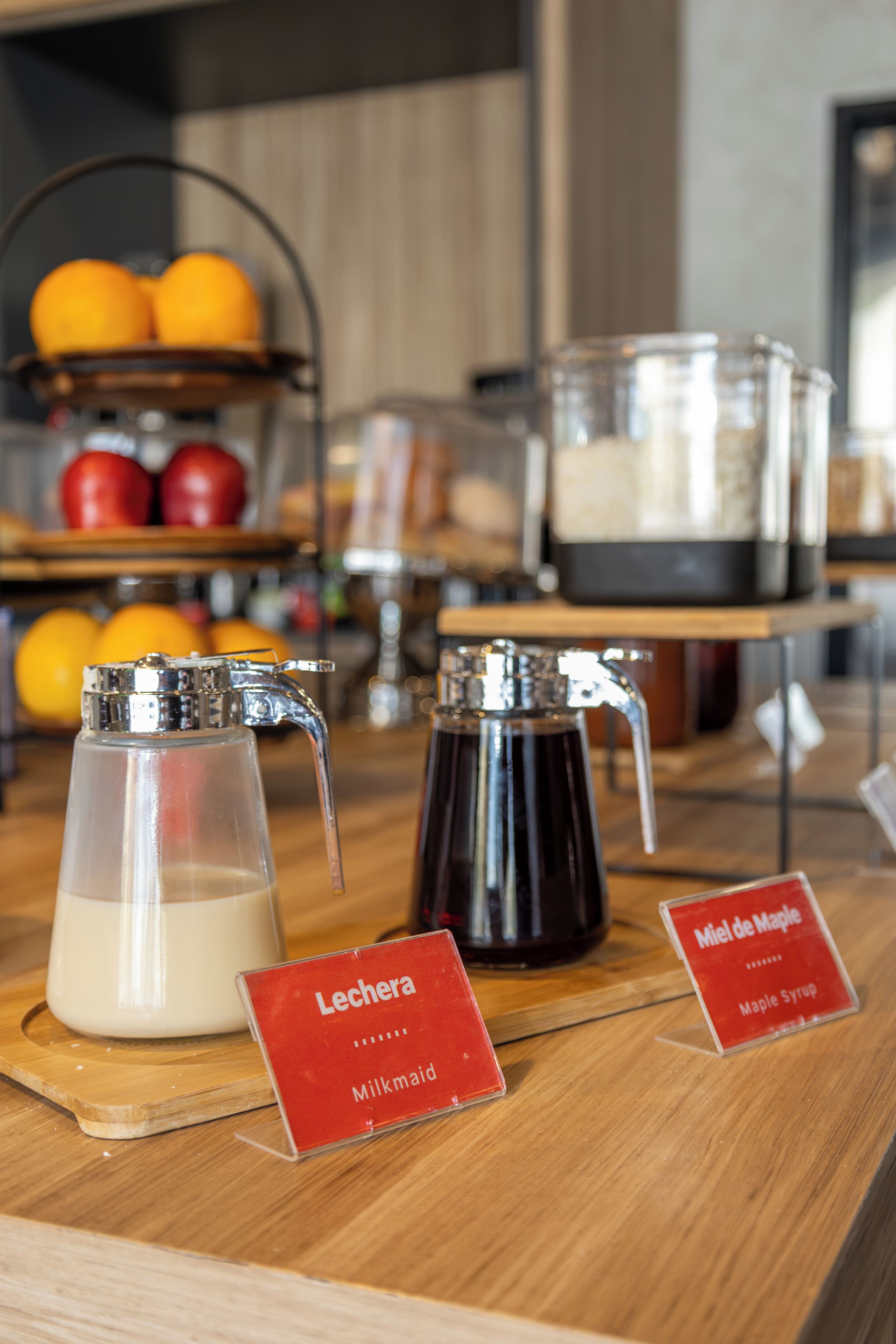 A coffee station with a milk dispenser and a syrup dispenser, along with labels for Lechera and Miel de Maple.