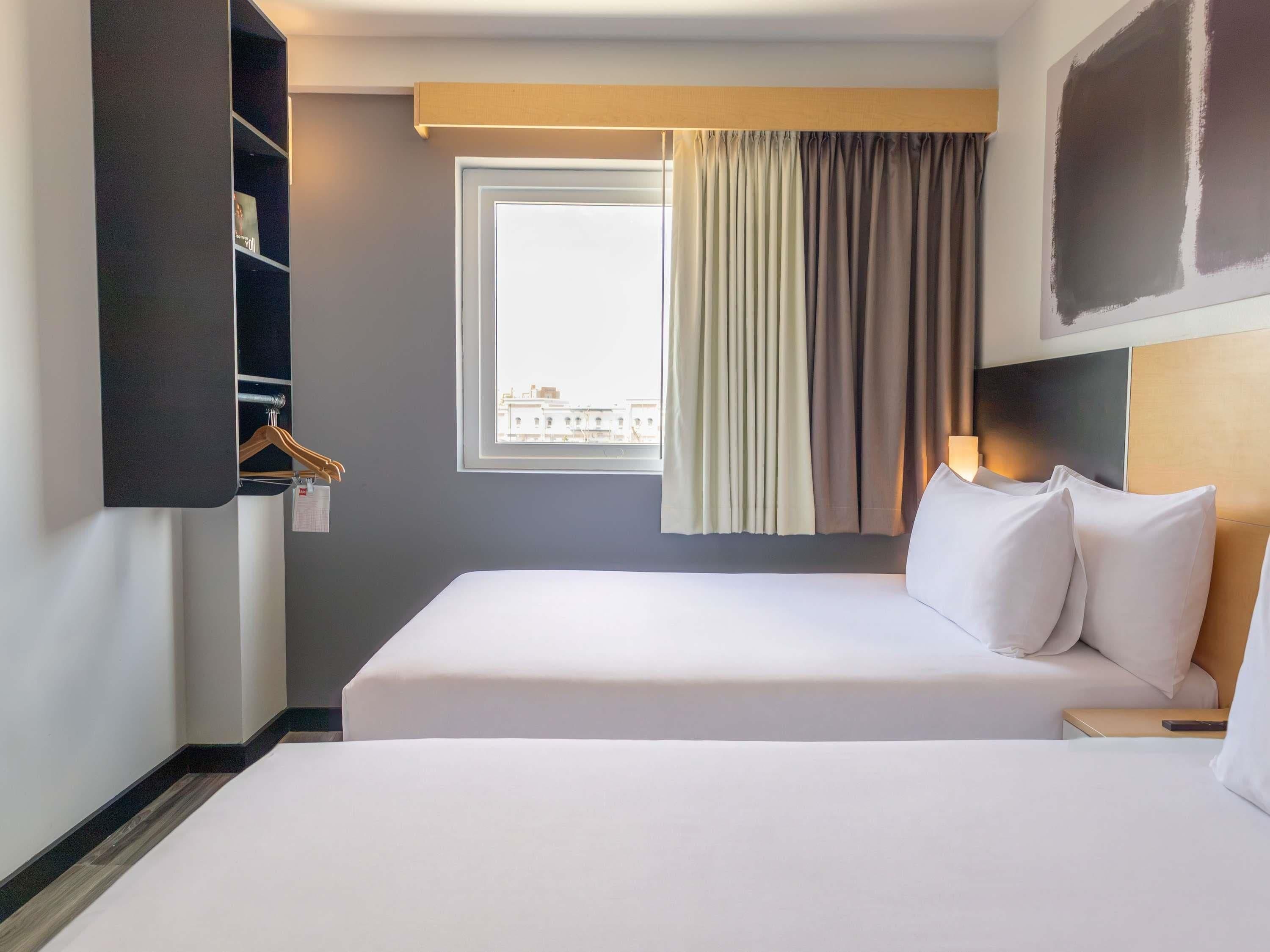 Una habitación de hotel con una cama grande, una mesita de noche, perchero y una ventana con cortinas que ofrece vistas a edificios.