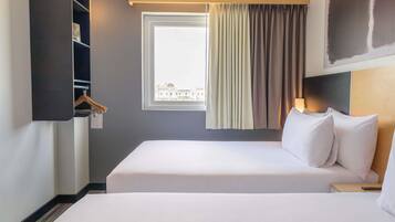 Una habitación de hotel con una cama grande, una mesita de noche, perchero y una ventana con cortinas que ofrece vistas a edificios.