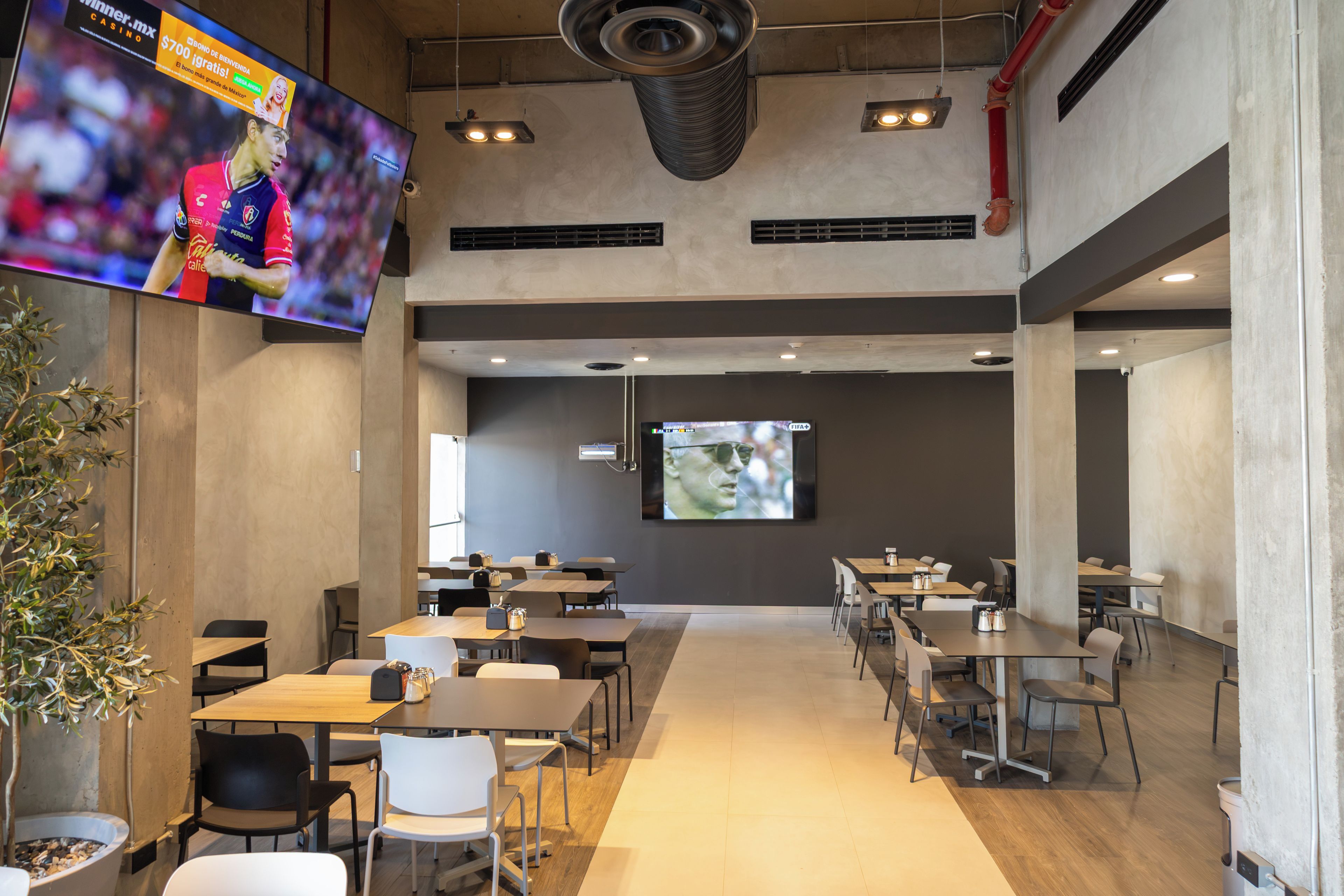 Un área de comedor moderna con varias mesas y sillas, una pantalla grande mostrando un evento deportivo y una planta en maceta.
