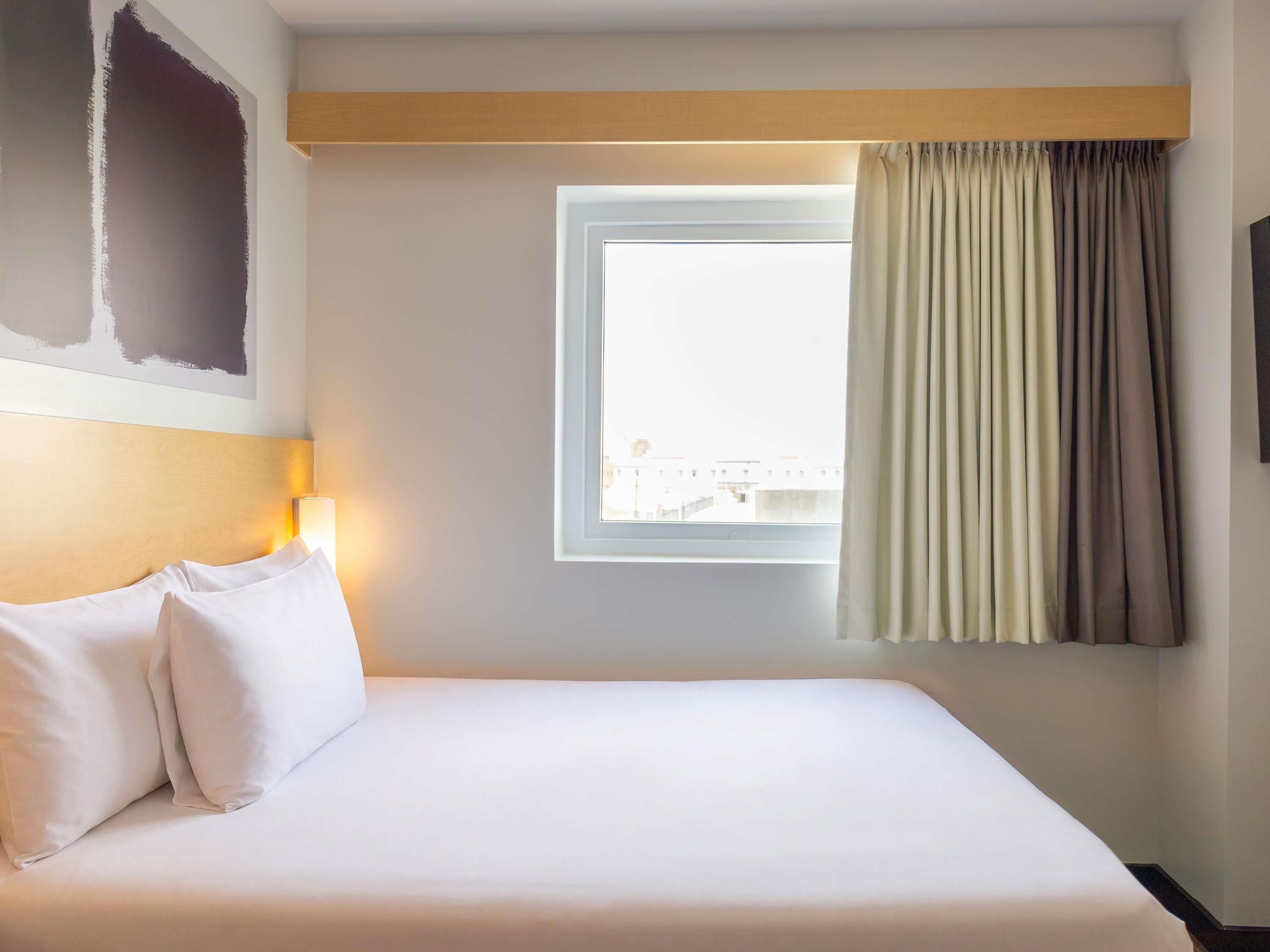 A hotel room with a bed, a window with curtains, a wall-mounted lamp, and a view of buildings outside.