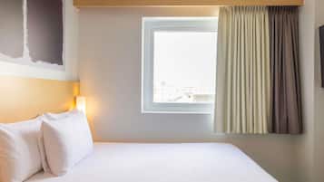 A hotel room with a bed, a window with curtains, a wall-mounted lamp, and a view of buildings outside.