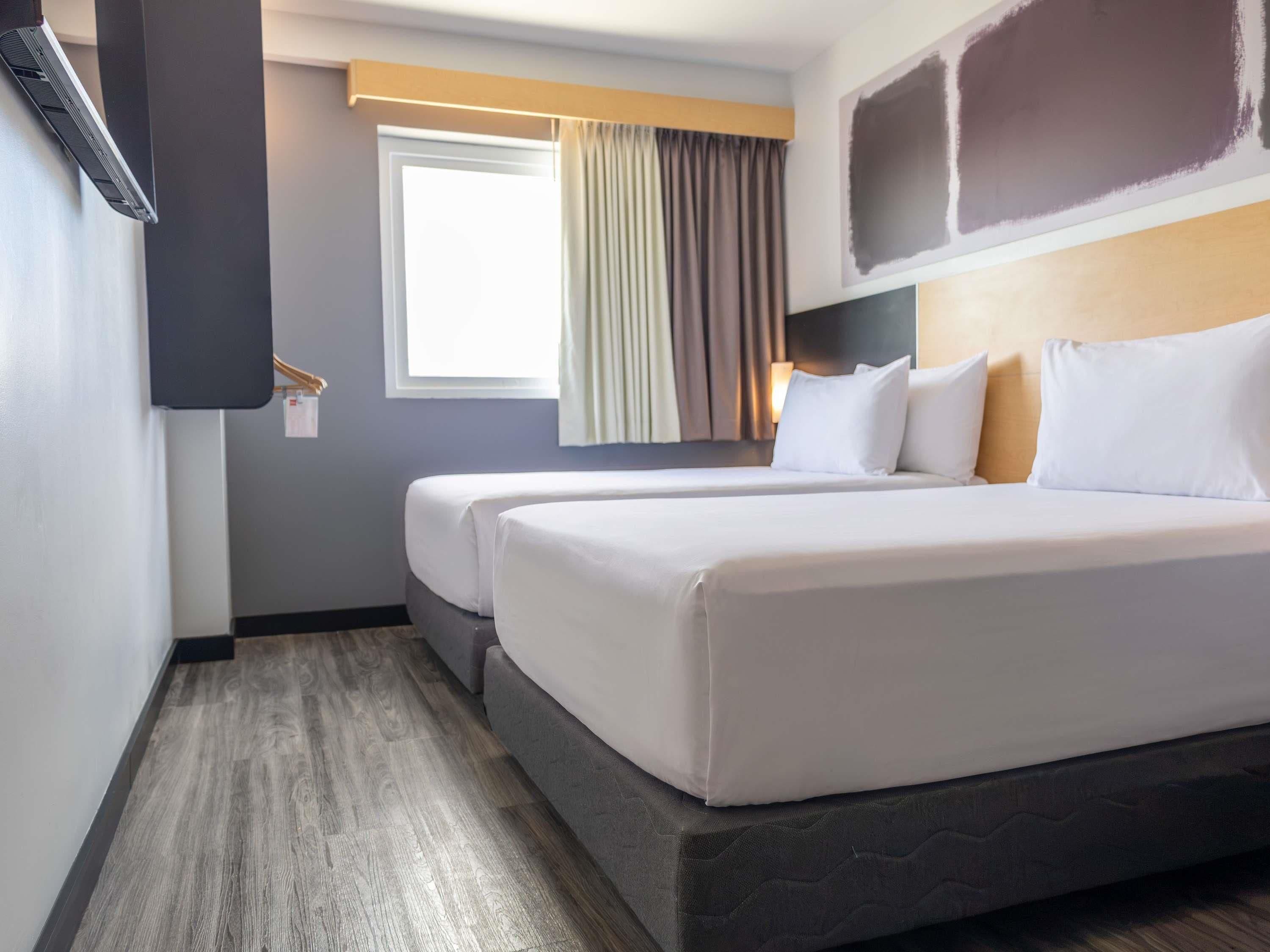 A hotel room with two beds, a window with curtains, a wall-mounted TV, and a wooden floor.