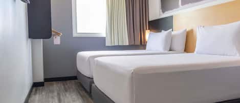 Una habitación de hotel con dos camas, un ventanal con cortinas, un televisor instalado en la pared y piso de madera.