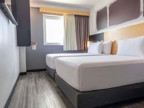 A hotel room with two beds, a window with curtains, a wall-mounted TV, and a wooden floor.