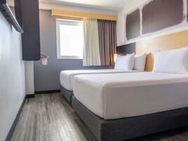 A hotel room with two beds, a window with curtains, a wall-mounted TV, and a wooden floor.