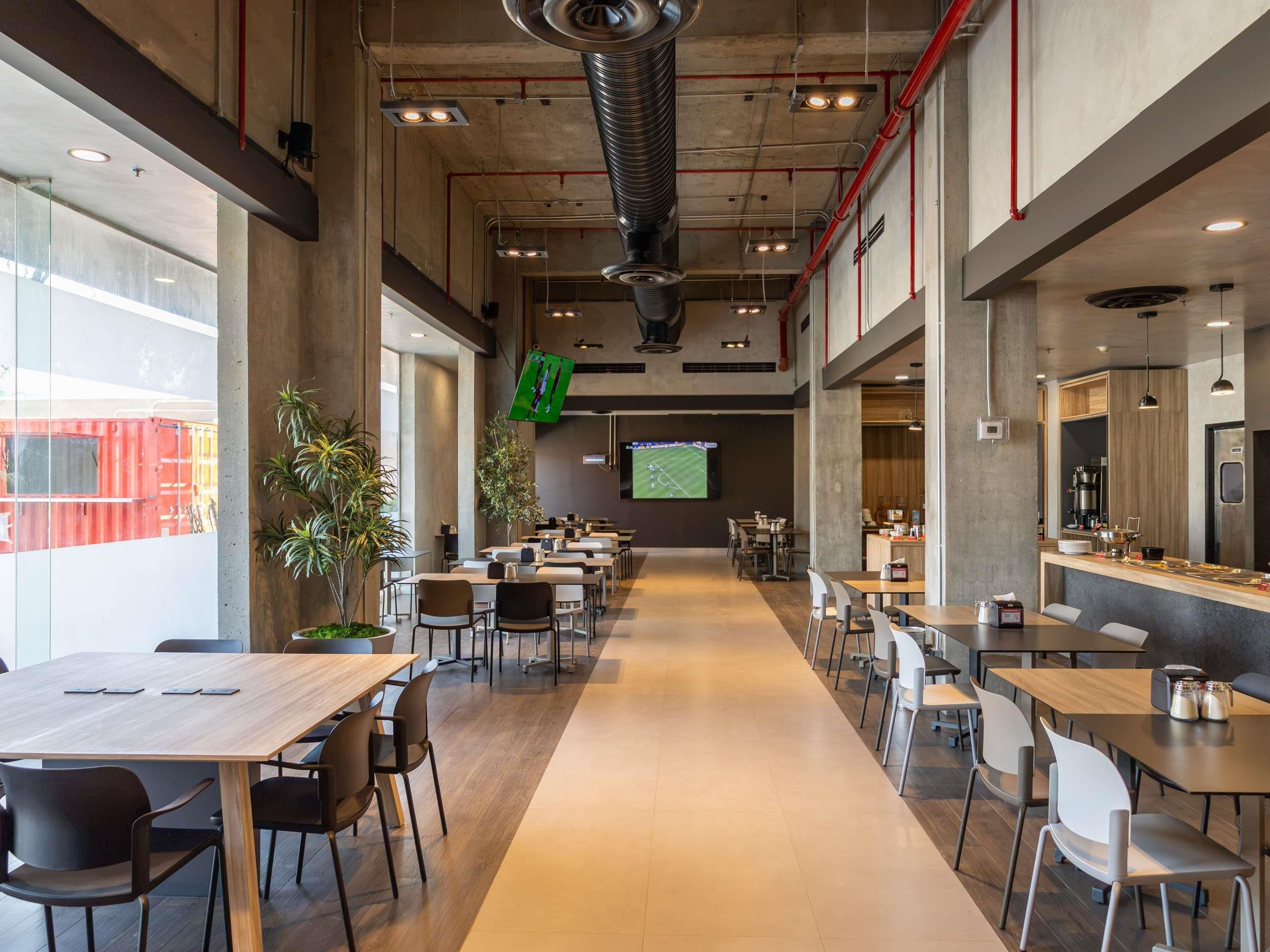 A spacious indoor dining area with multiple tables and chairs, a large screen displaying a sports event, and a kitchen area with modern appliances.