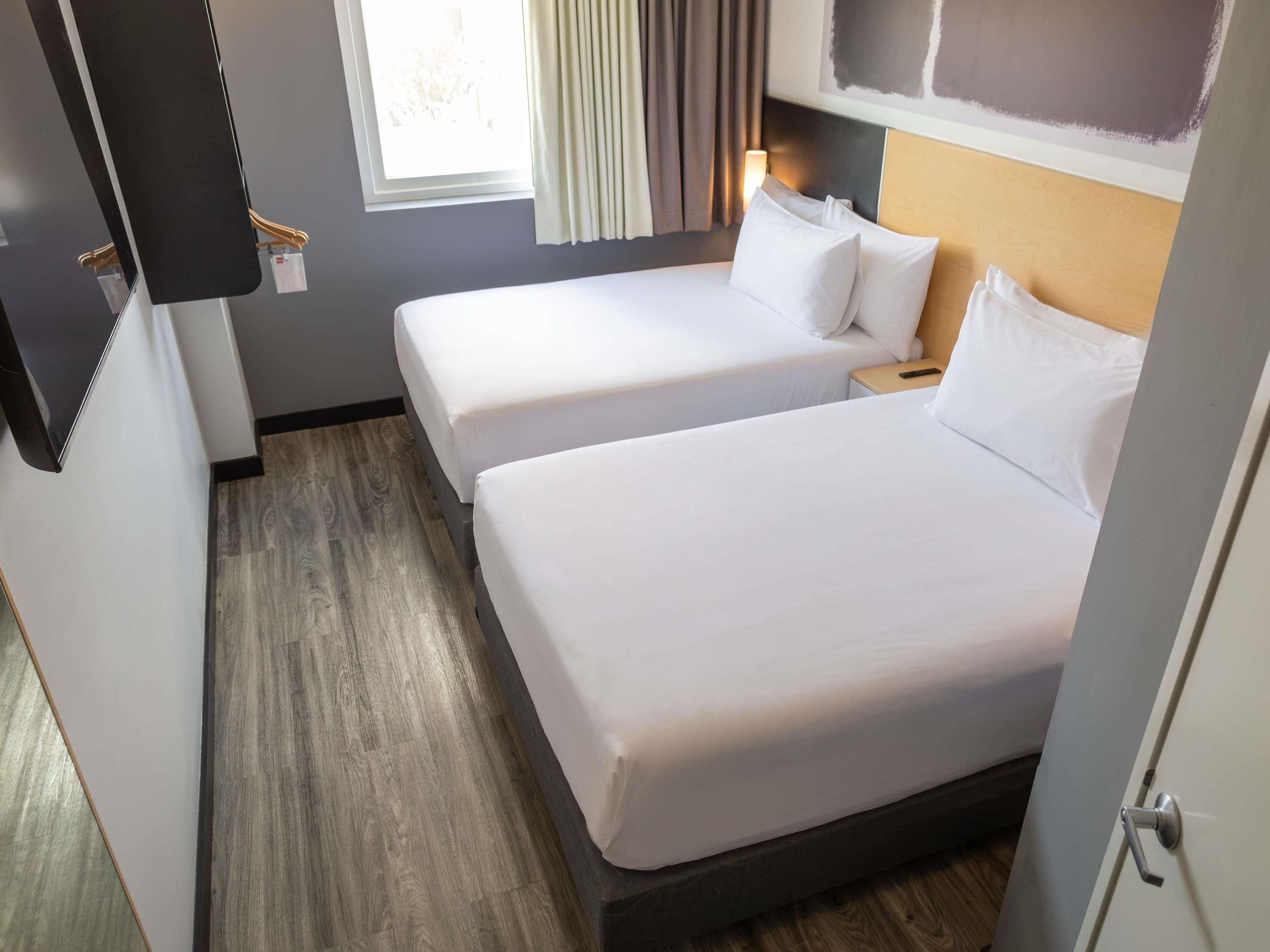 Habitación de hotel con dos camas individuales, suelo de madera, una ventana con cortinas y un televisor montado en la pared.