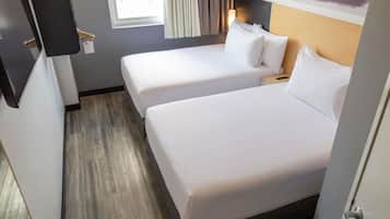 Habitación de hotel con dos camas individuales, suelo de madera, una ventana con cortinas y un televisor montado en la pared.
