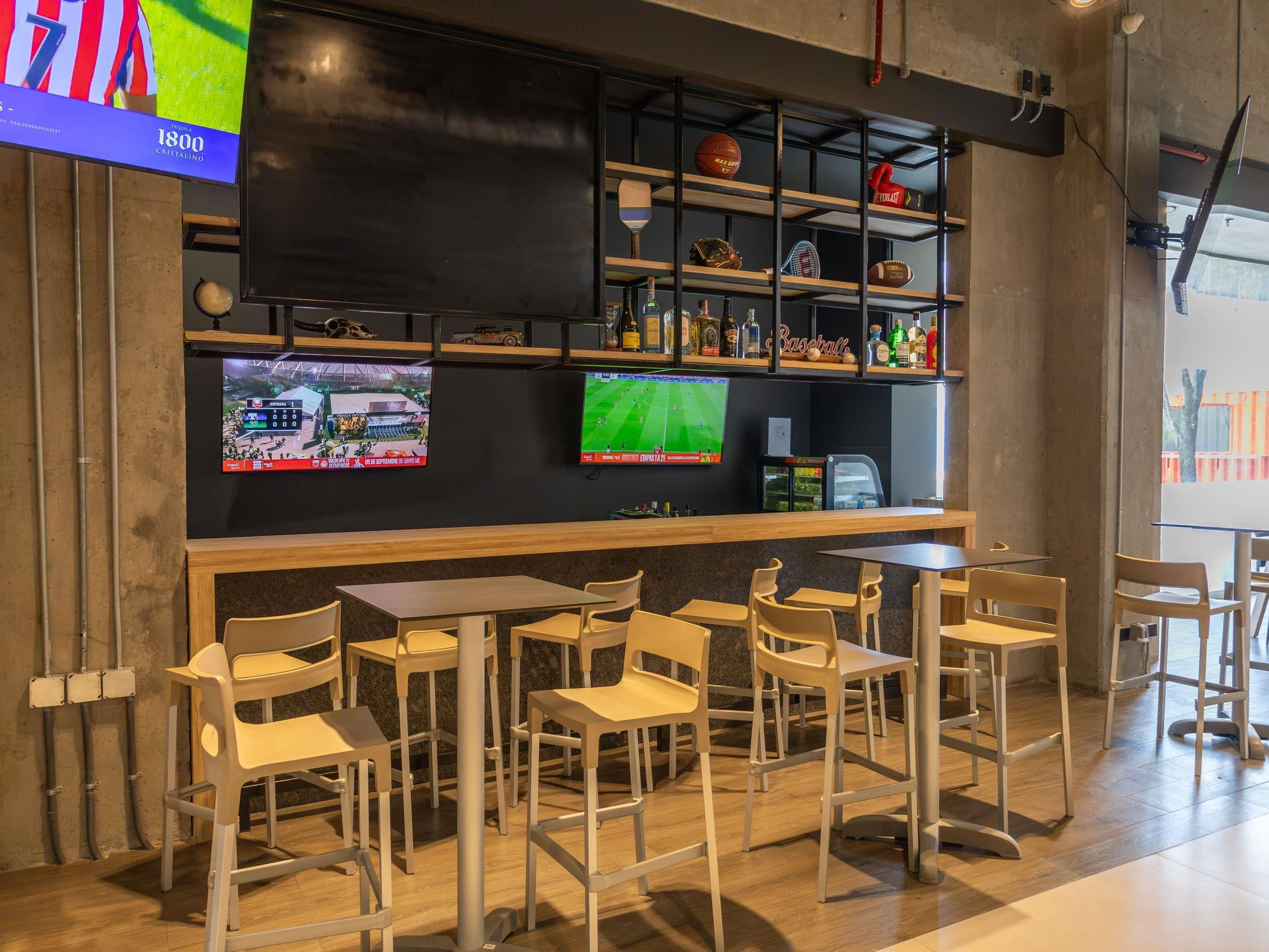 Zona de bar con diferentes opciones de asientos, un televisor mostrando deportes y estanterías con diversos artículos.