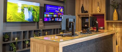 A reception area with a wooden reception desk, a flat-screen TV displaying a nature scene, a digital screen with various icons, and shelves with decorative items.