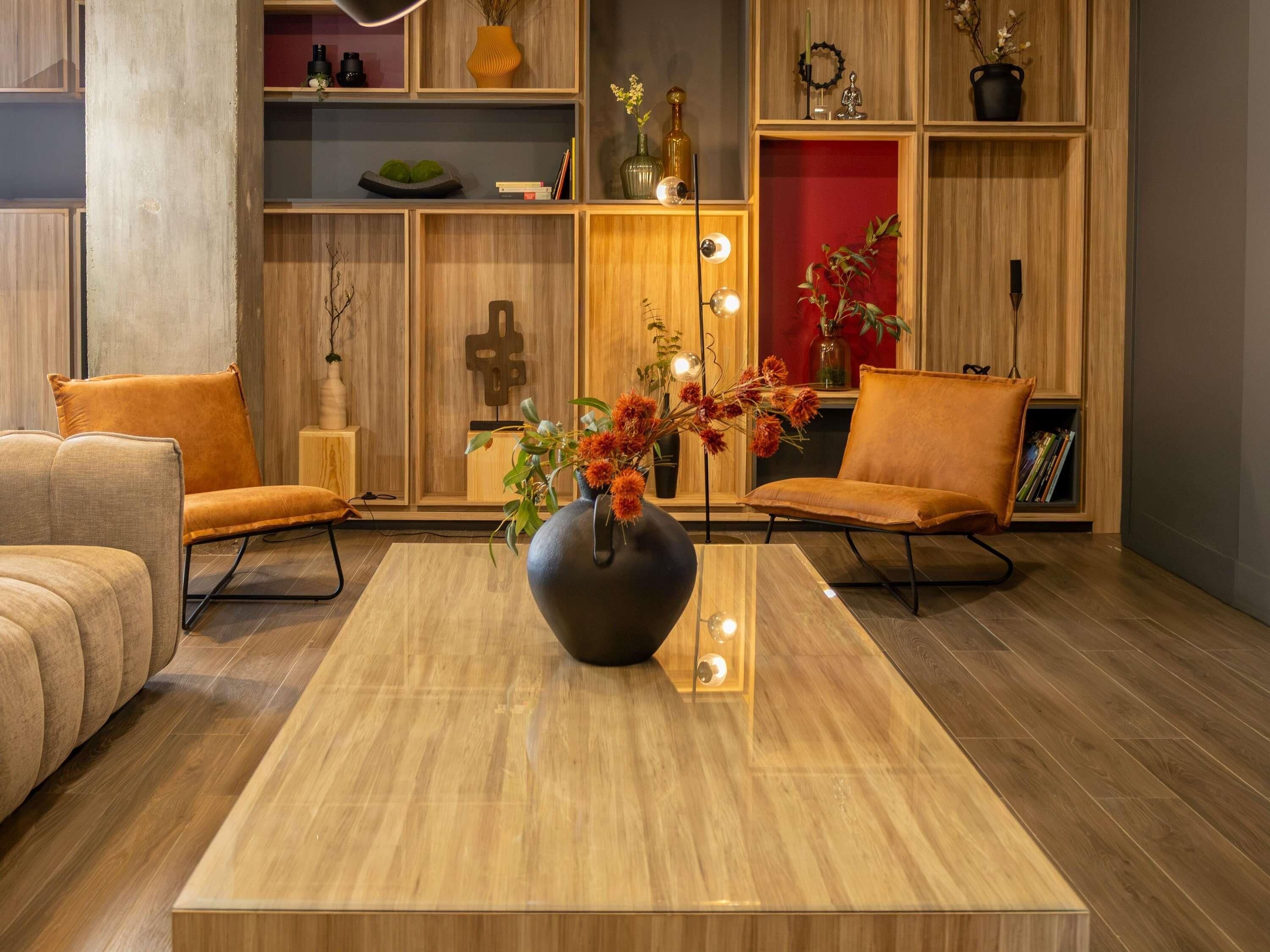 A modern living room with a wooden floor, two orange armchairs, a central coffee table with a vase, and wooden shelving with various decorative items.